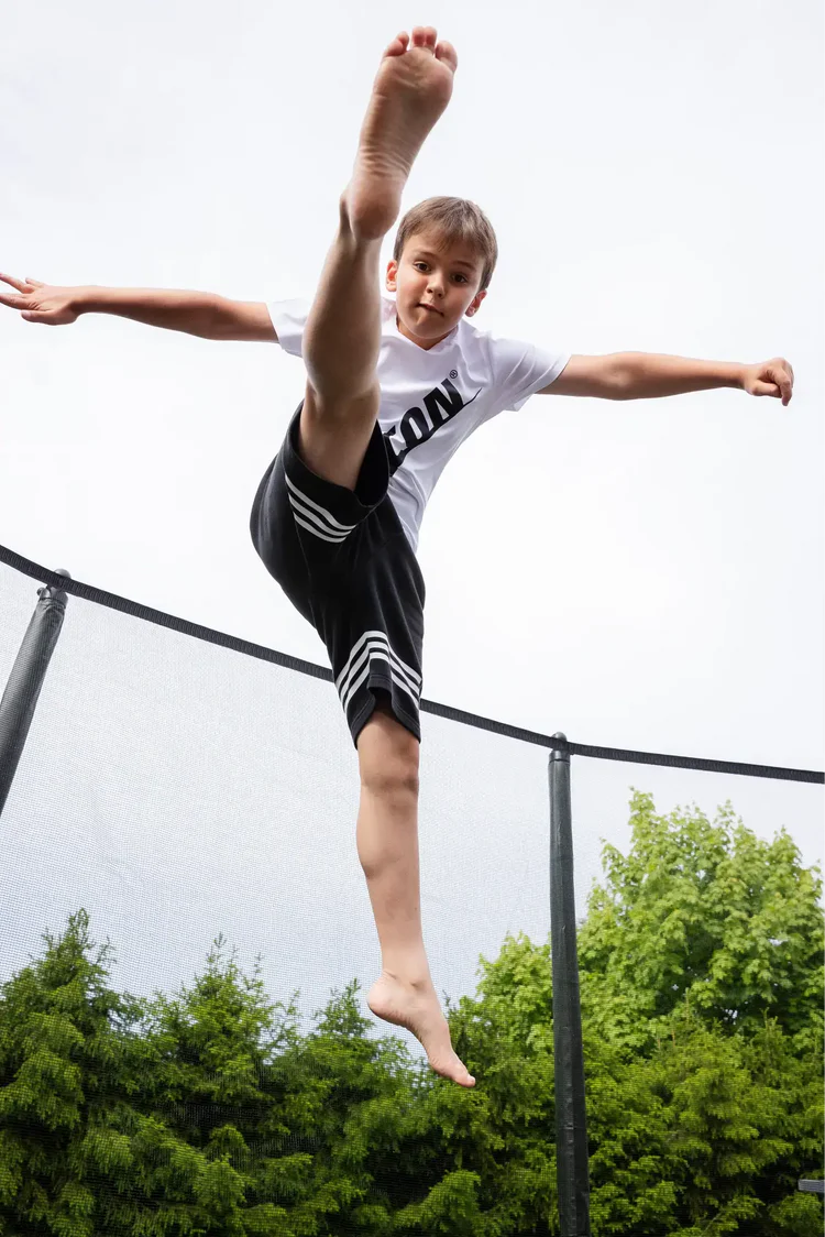 A child wearing a white Acon t-shirt performing a high kick on a trampoline, with one leg extended above his head.