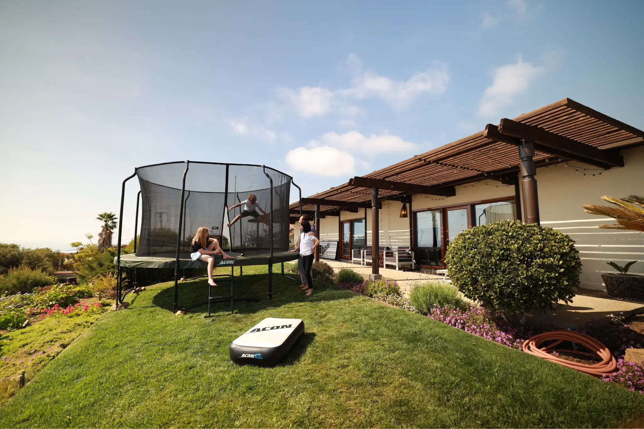 A group of children with their parents enjoying an Acon trampoline in a sunny backyard with a modern house, green lawn, and garden in the background.