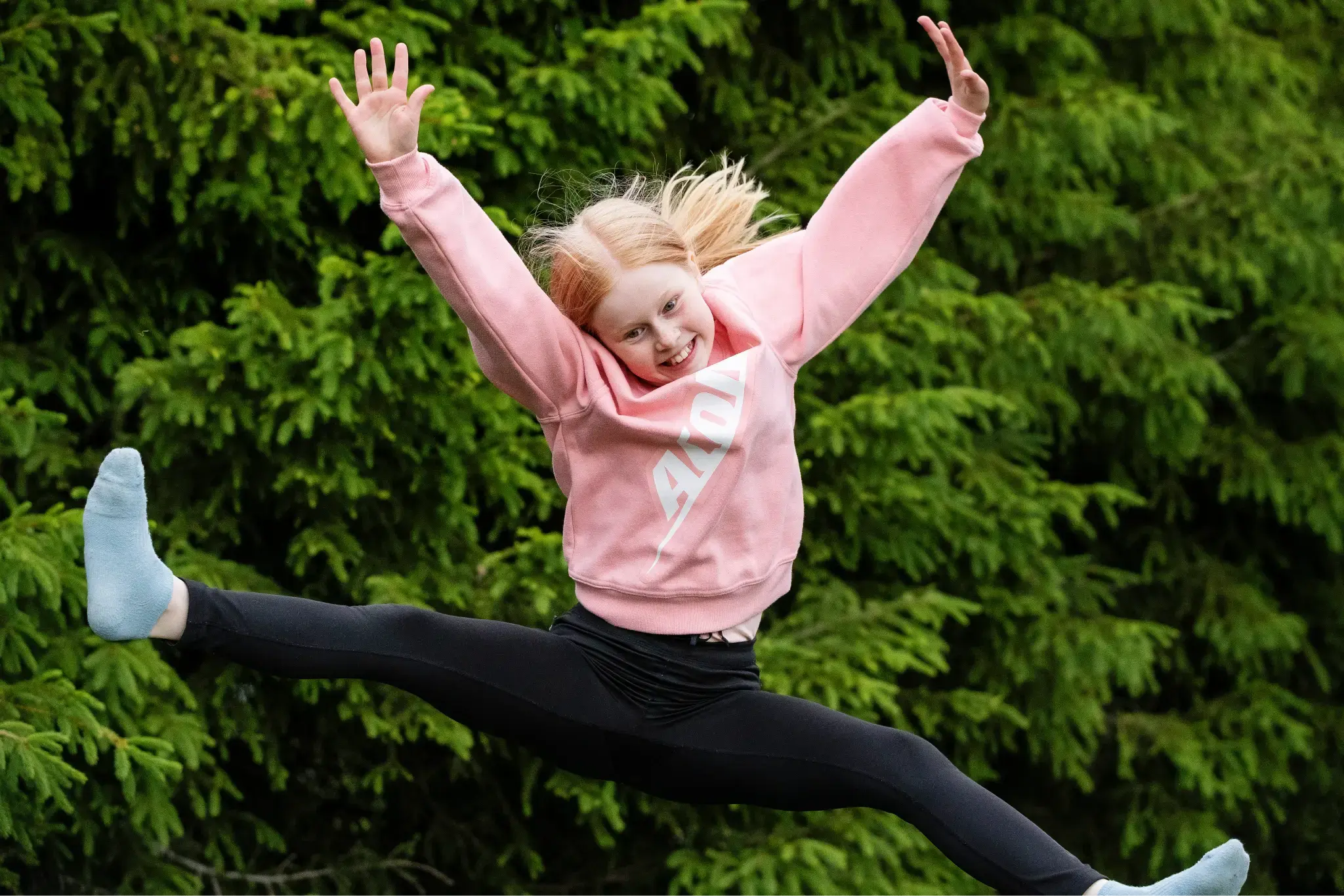 A young girl in a pink Acon sweatshirt jumps on a trampoline with arms up and legs in a split. Green trees fill the background.