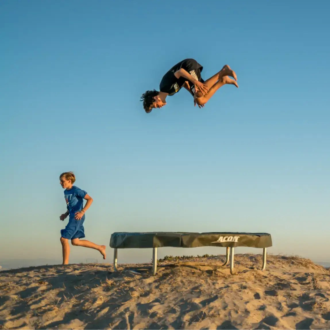 Two children playing on a trampoline set against a sandy beach backdrop at sunset. One performs a backflip while the other runs nearby