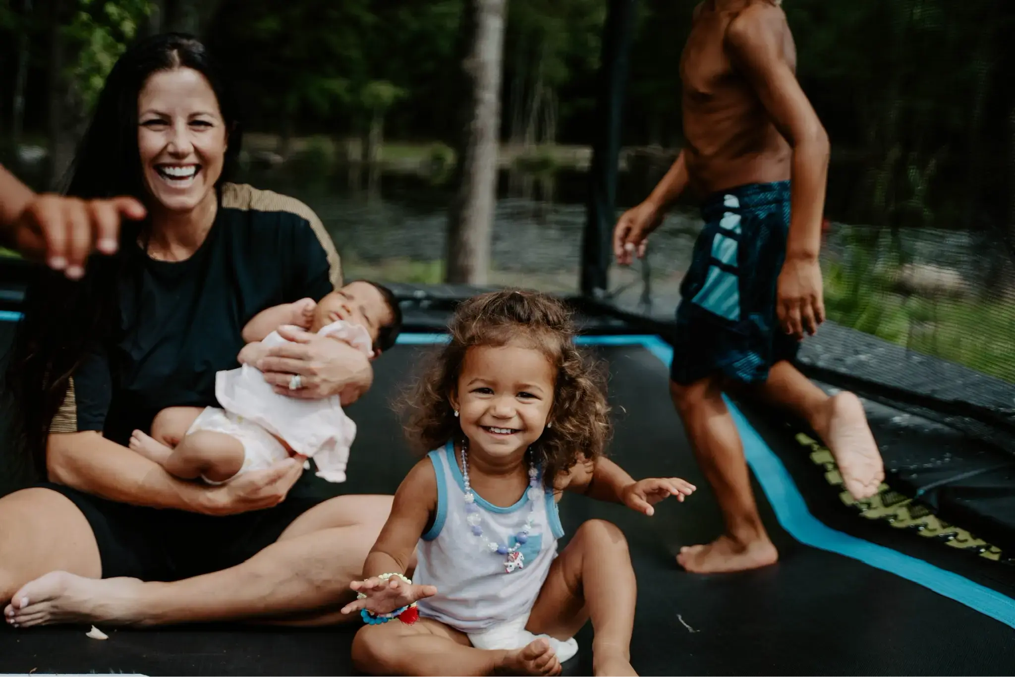 A smiling mother holds a baby while sitting on a trampoline, surrounded by happy children playing and enjoying a fun outdoor moment.