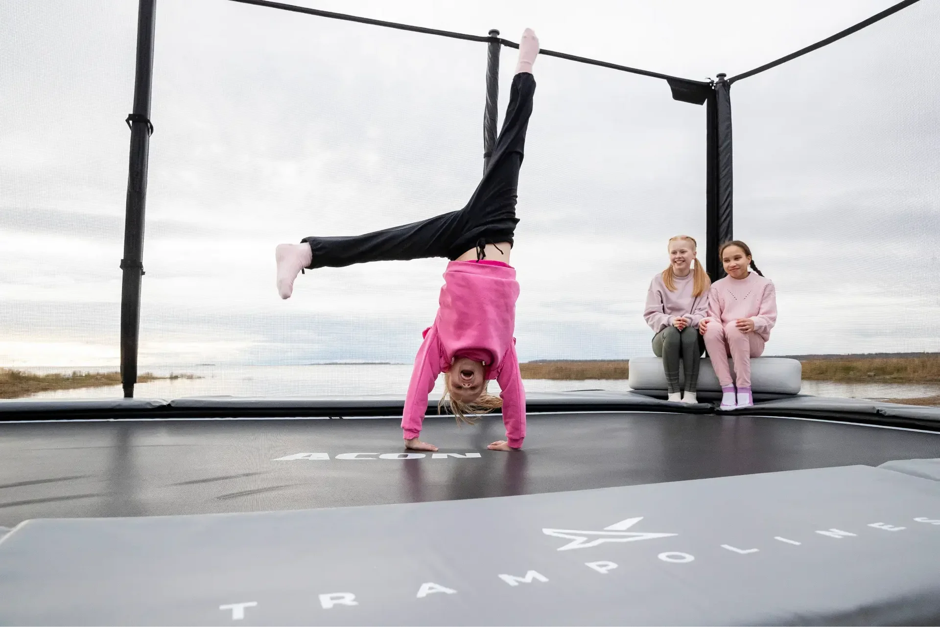 Girl doing a handstand on an Acon X trampoline, with two others sitting nearby.