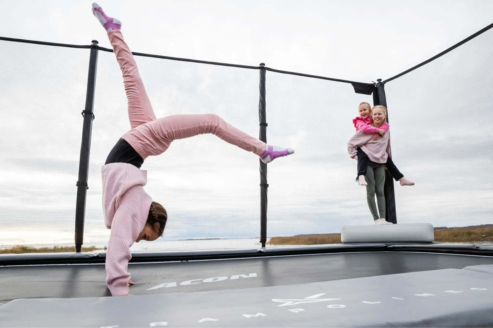 Three children enjoying a trampoline. One performs a handstand, while another carries a younger sibling, set against a serene outdoor backdrop.