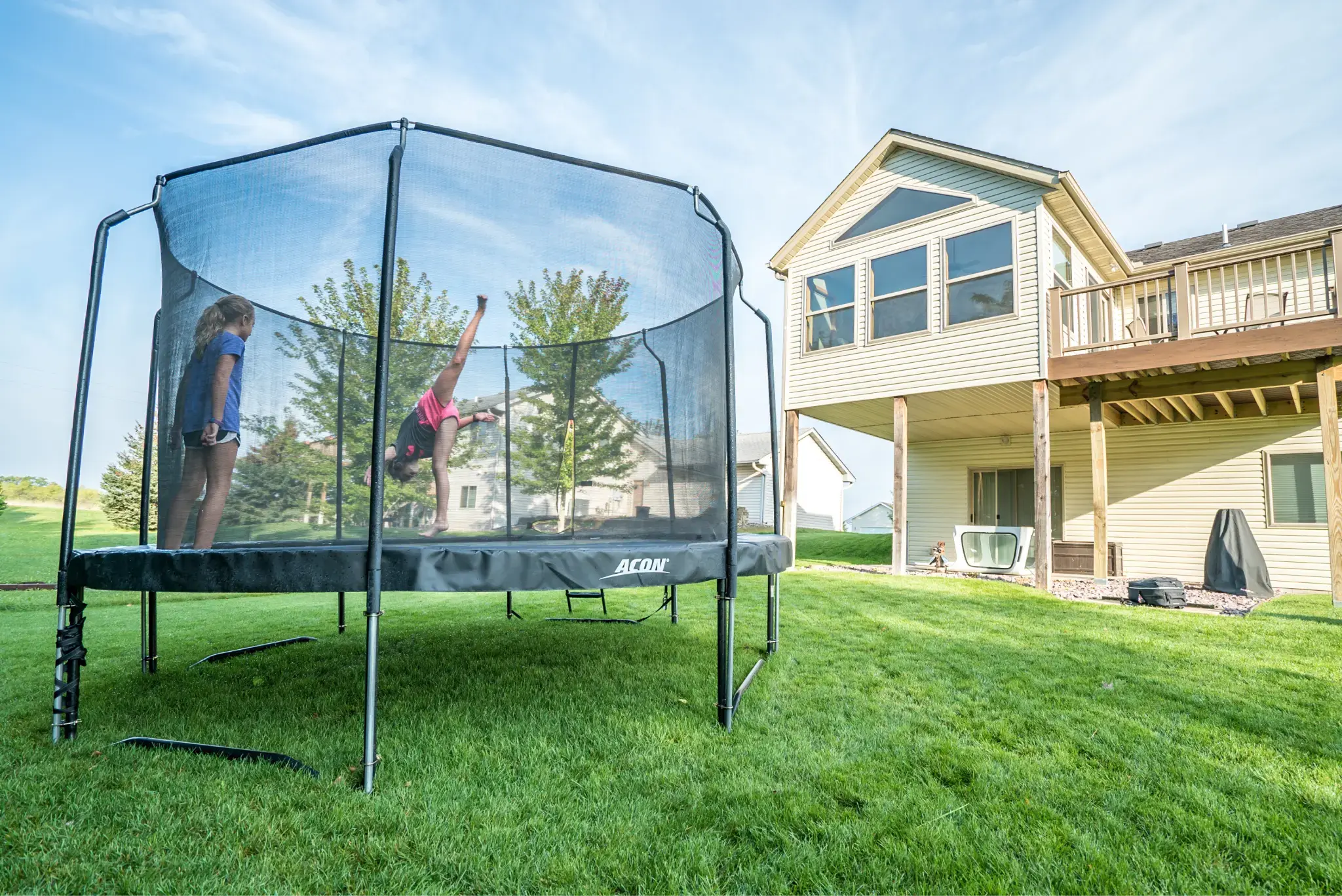 Two children playing on a large Acon trampoline with a safety net in a sunny backyard, surrounded by green grass and a modern house.