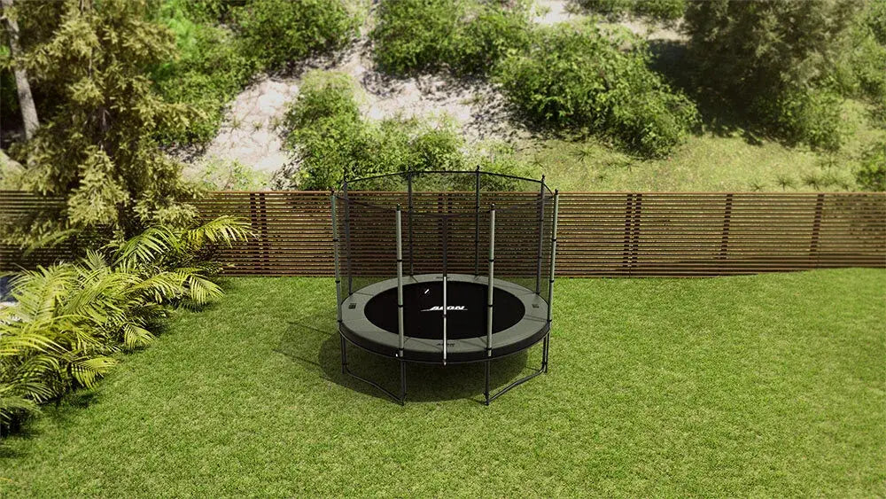 A round Acon trampoline with a safety net is set up in a backyard with green grass, a wooden fence, and lush plants in the background.