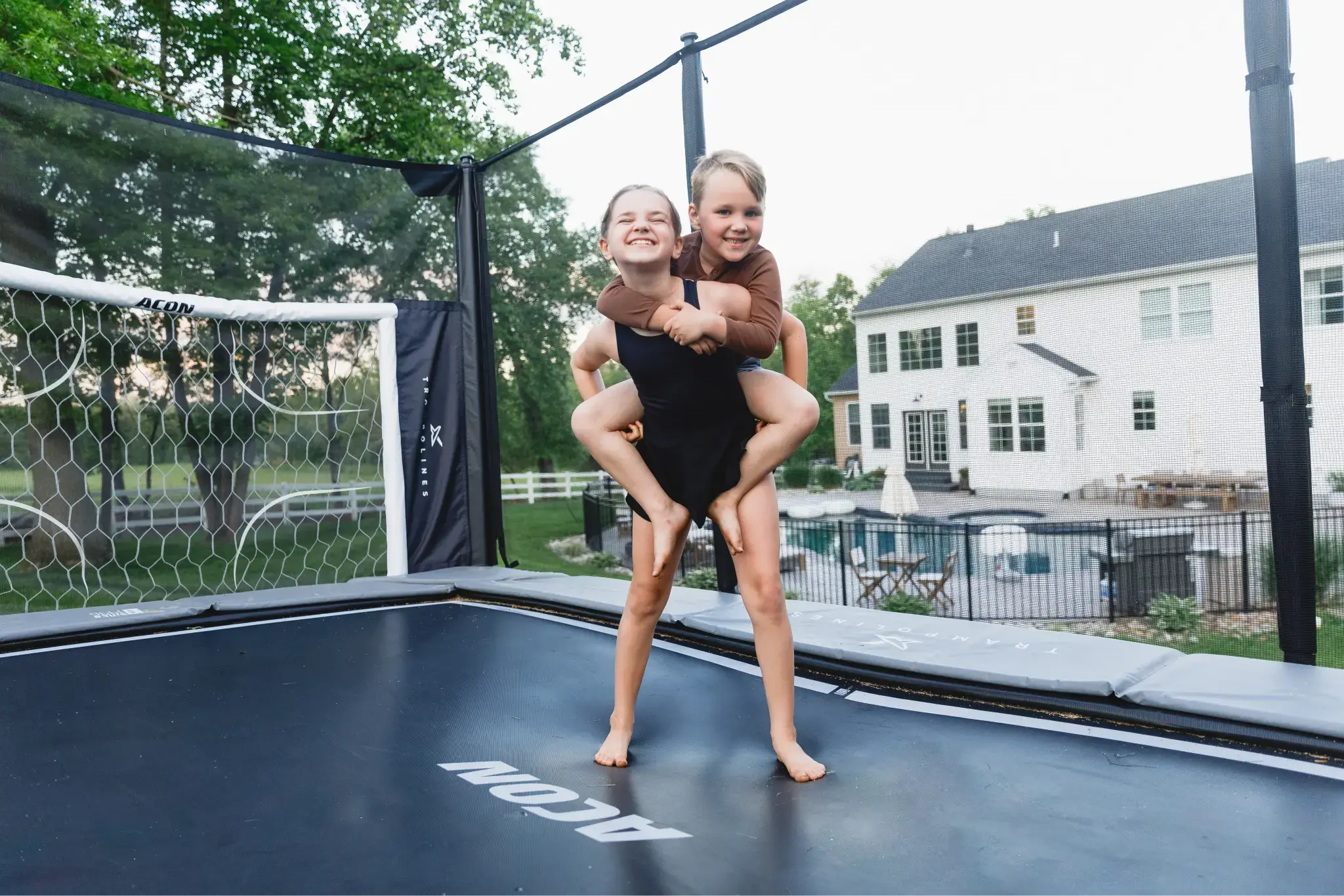 Two kids smiling and playing on a trampoline, one giving the other a piggyback ride, with ACON X soccer goal panel in the background.