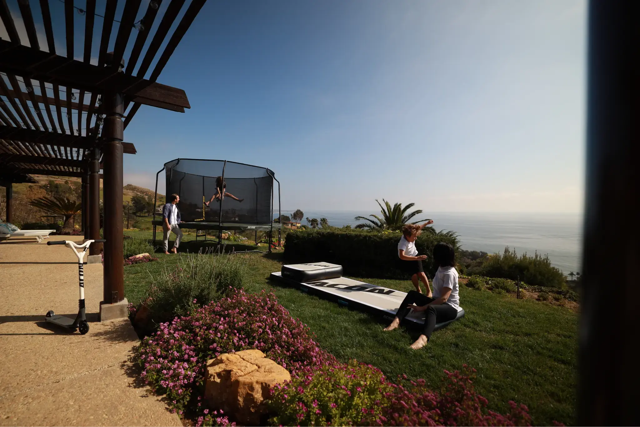 Family enjoying a sunny backyard by the ocean, equipped with a trampoline and an airtrack, surrounded by greenery.