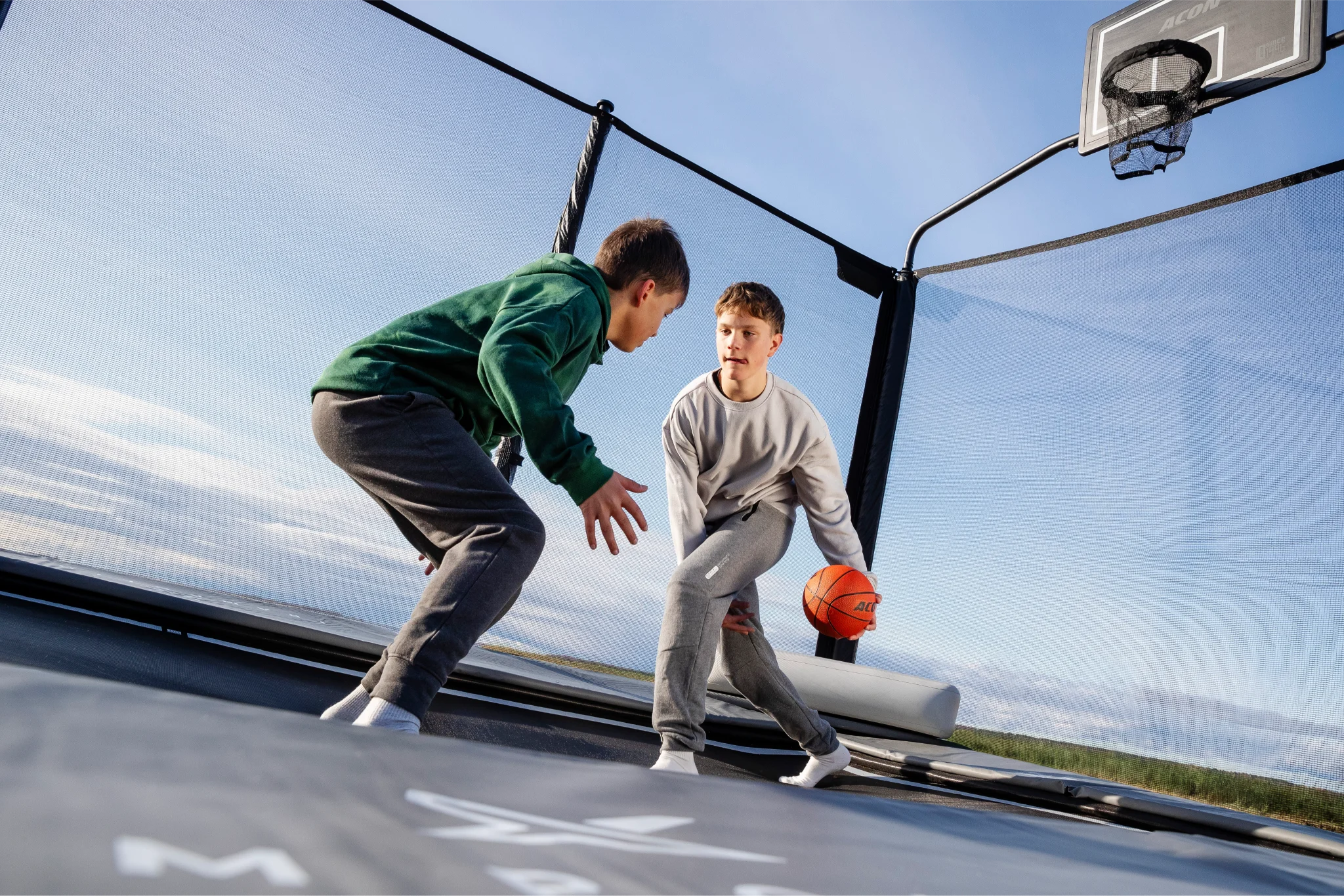 Two teenagers playing basketball on an Acon trampoline with a basketball hoop.