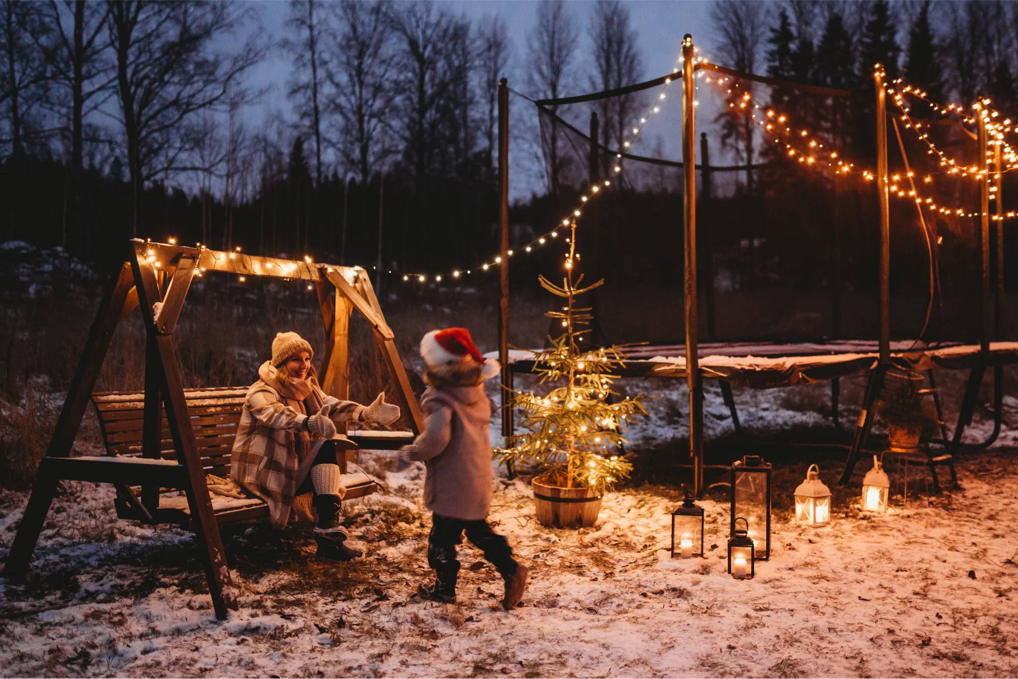 Child going to their mother on who is sitting on a swing.  Winter night with snow on the ground, and an Acon trampoline with Christmas lighting on the background.