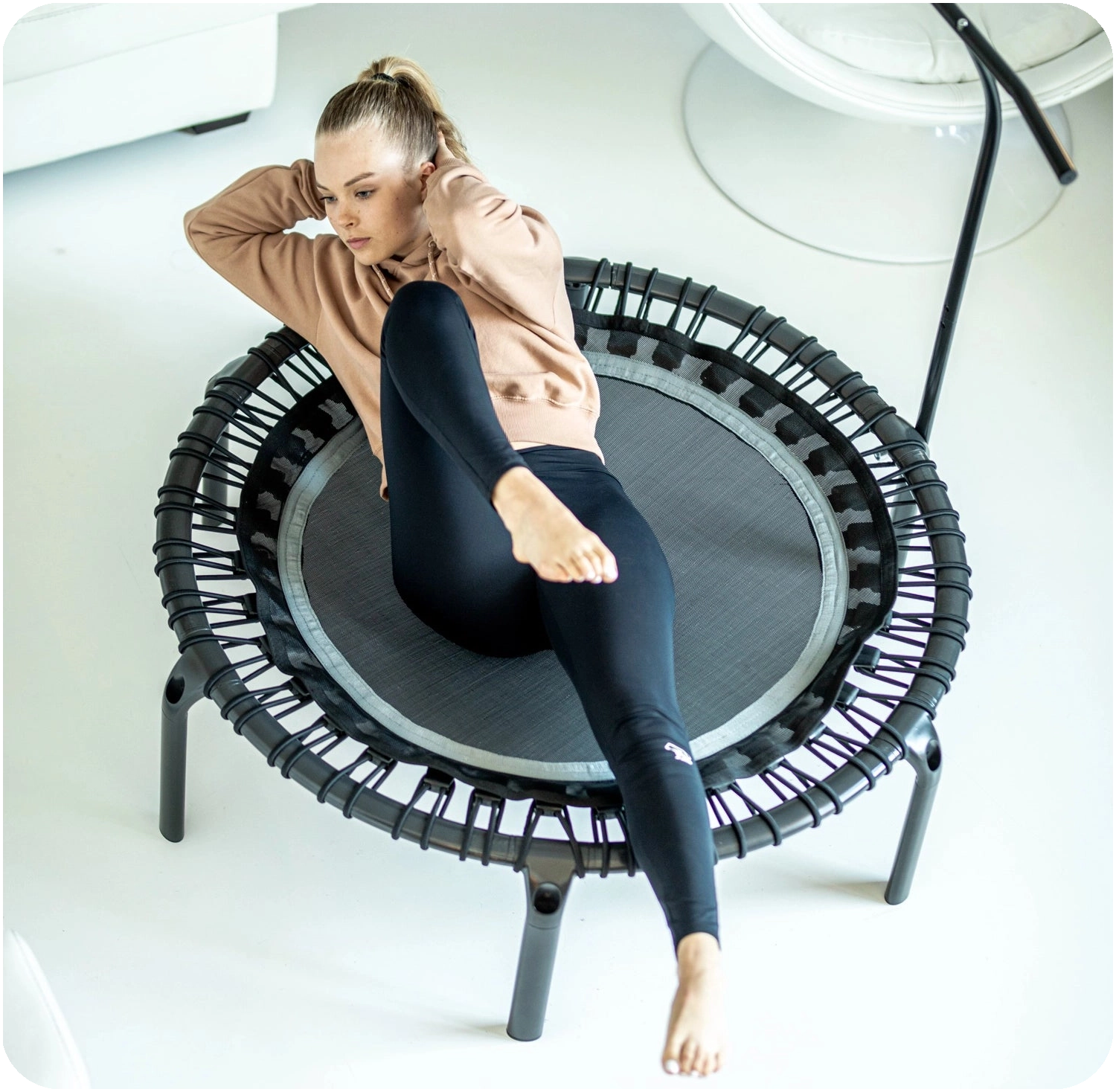 A woman gymnasts on a black round Acon FIT rebounder in a Nordic-style living room.