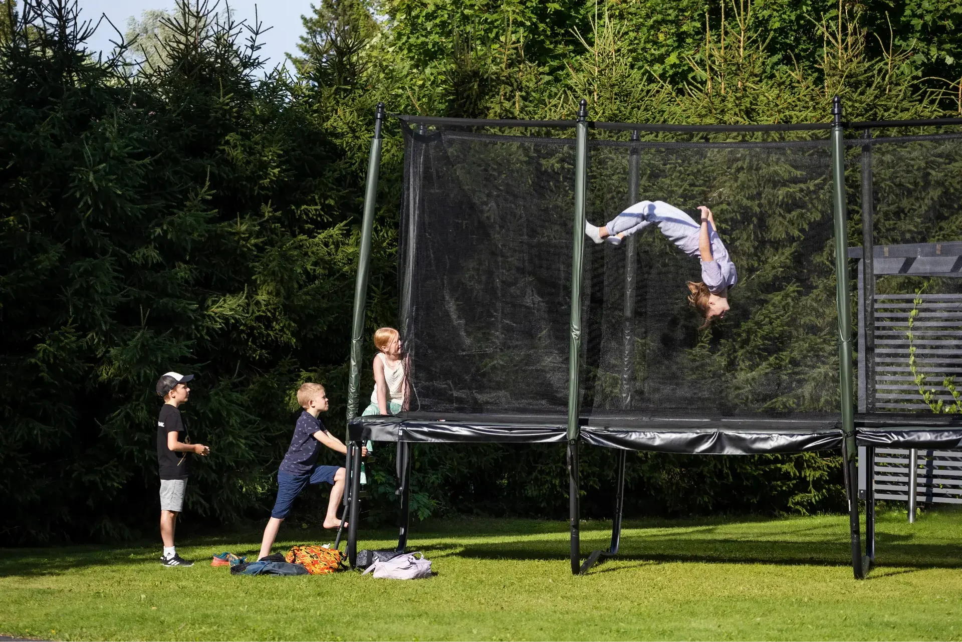 Group of kids playing on a trampoline with a safety net outdoors, with one child flipping mid-air and others waiting their turn.