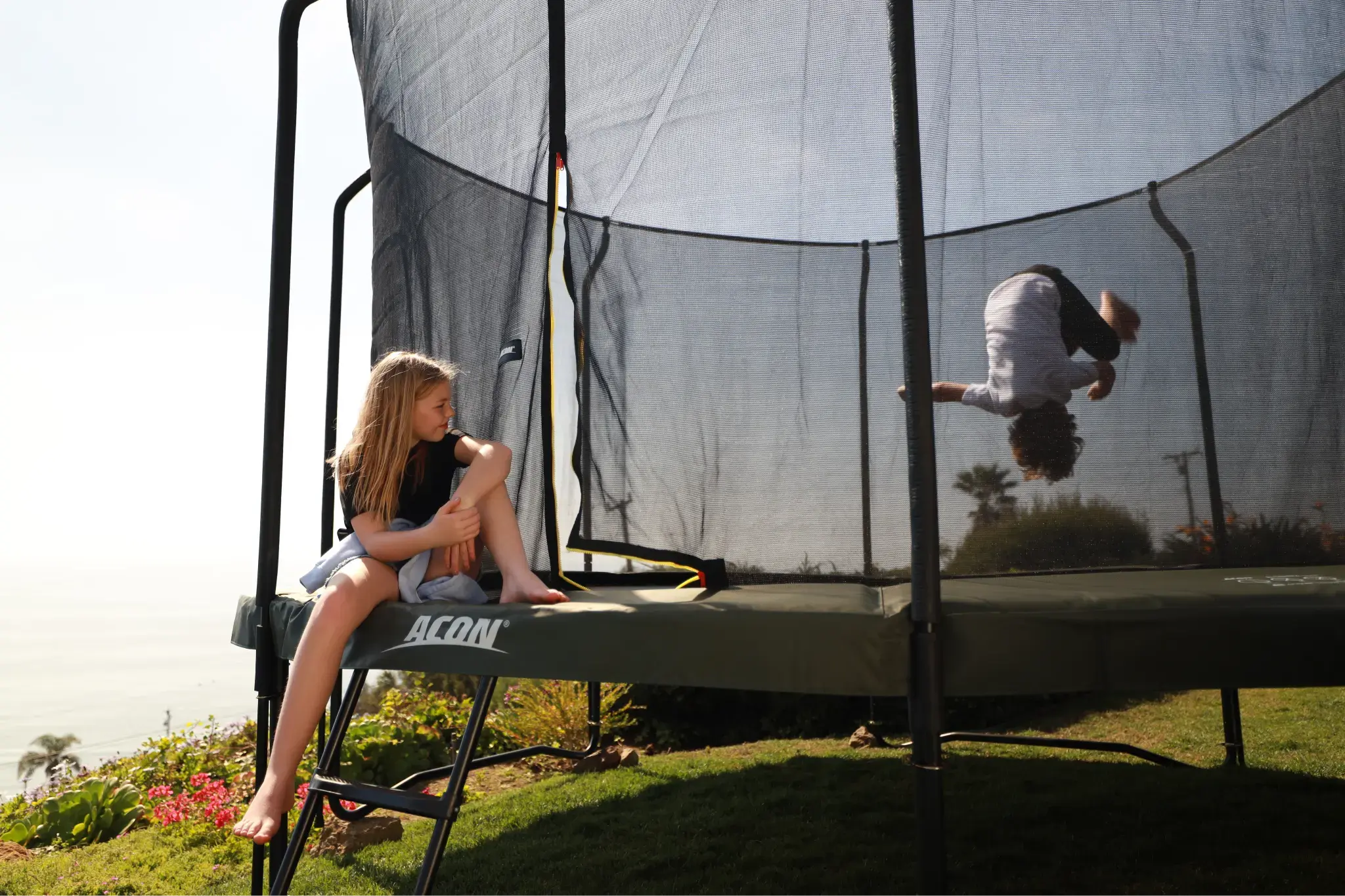 A girl sits on the edge of an Acon trampoline while another child does a flip inside the safety net. The background features a sunny outdoor setting with plants and ocean views.