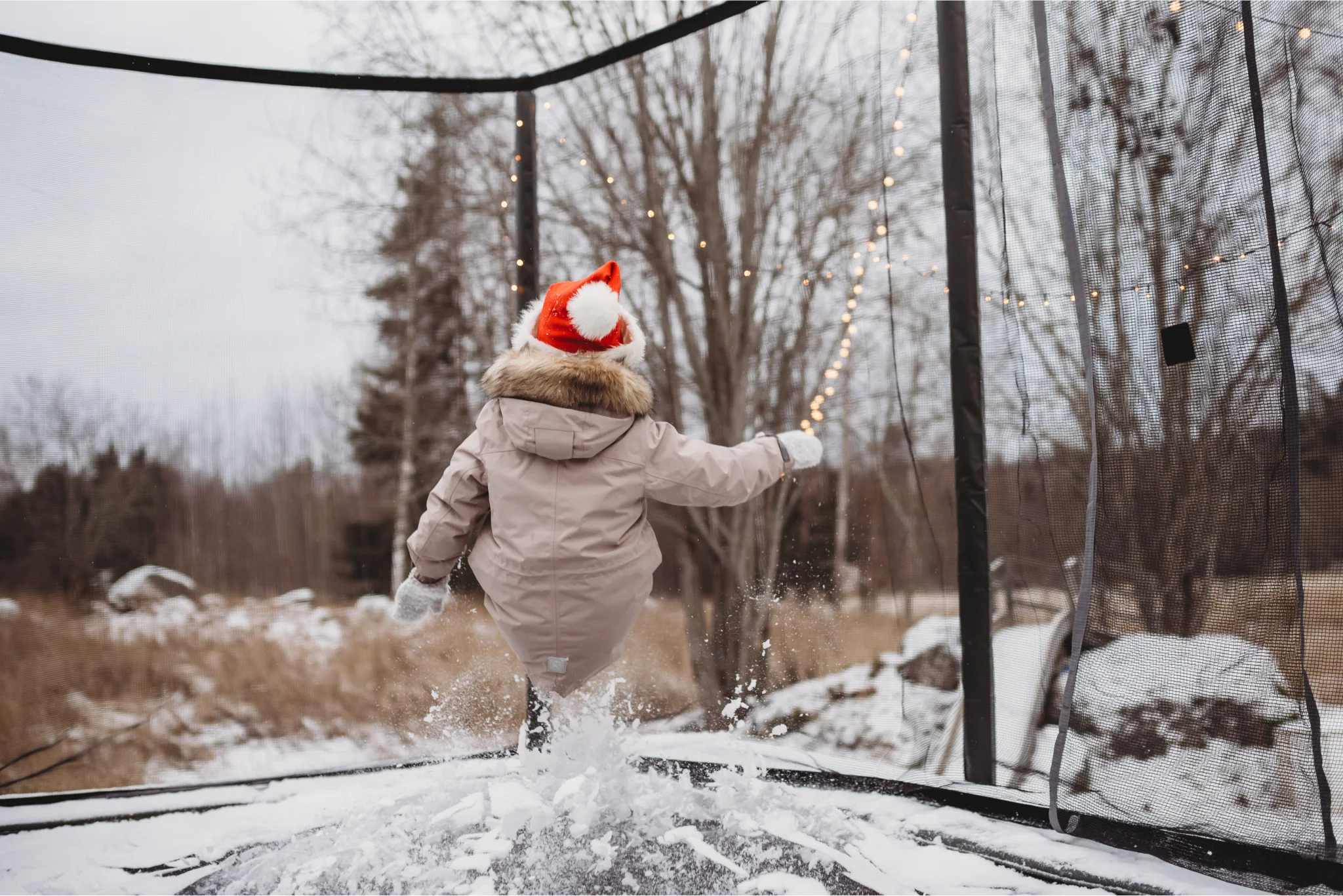 Small child with a winter coat and a santa hat jumping on a trampoline with snow.