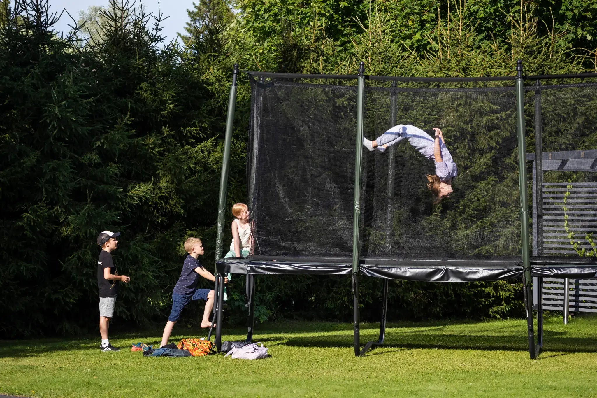 A girl doing a backflip on a garden trampoline with a safety net while other children are waiting their turn.